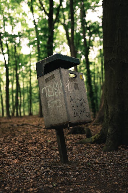A weathered grey metal post box with a slightly open black lid, mounted on a tilted metal pole, standing amidst a woodland area with tall trees and dense green foliage in the background. The post box surface is covered in graffiti tags and markings, with some faded paint that suggests exposure to the elements over time. The ground around it consists of compacted earth and scattered leaves, indicating a forest trail or path. The lighting is natural, with diffused sunlight filtering through the tree canopy, creating a soft, shaded environment. This scene captures a quiet, outdoor setting where the post box appears out of place among the natural surroundings, reflecting a form of independent waste or item disposal, akin to alternative rubbish handling outside conventional urban waste collection points.