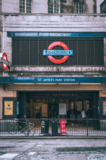 The entrance to St. James's Park Station, part of the London Underground, is visible in the image, featuring a blue sign with white lettering displaying the station name. Above the entrance, there is a large circular underground logo with a red ring and a blue horizontal bar with the word 'UNDERGROUND' in white letters. The facade of the station shows signs of weathering and grime, with dark streaks and dirt around the signage and metal grille. In front of the entrance, a black metal railing runs parallel to the sidewalk, helping to organize pedestrian flow, and a silver bicycle is parked against it on the left side of the image. To the right of the bicycle, there is a small orange and black litter bin, and a red and white A-board sign placed on the pavement, possibly advertising nearby services or events. The street in front appears wet, likely from recent rain, with reflections visible on the asphalt surface. The environment suggests an urban setting in central London, where private waste collection services might be employed for waste disposal related to station staff or nearby establishments, complementing the usual municipal rubbish removal. The scene is illuminated by natural daylight, contributing to the clear visibility of all elements, and the background shows the upper part of the station building with concrete and glass windows.