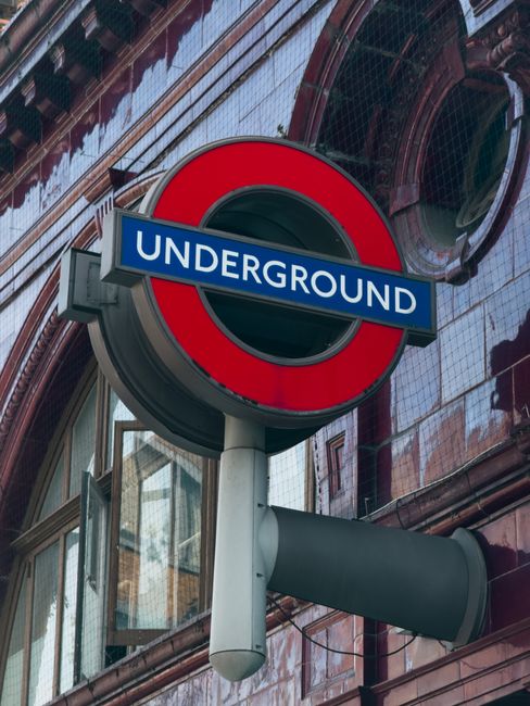 A close-up image of a London Underground station sign mounted on a metal pole in front of a building with reflective glass windows and dark red brickwork. The sign features a circular red border with a blue rectangle across the middle displaying white uppercase letters spelling 'UNDERGROUND.' The metal pole supporting the sign is painted light grey, extending horizontally with a partly visible cylindrical attachment beneath the sign. The background includes the reflections of nearby windows and some architectural details on the building's façade, with ambient daylight illuminating the scene. The setting suggests an urban environment, typical of underground station entrances, with an emphasis on clean, well-maintained signage that supersedes the scene's potential association with infrastructure or cityscape relevant to waste management and disposal context.