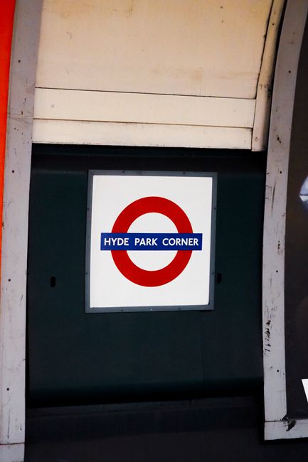 The entrance to St. James's Park Station, part of the London Underground, is visible in the image, featuring a blue sign with white lettering displaying the station name. Above the entrance, there is a large circular underground logo with a red ring and a blue horizontal bar with the word 'UNDERGROUND' in white letters. The facade of the station shows signs of weathering and grime, with dark streaks and dirt around the signage and metal grille. In front of the entrance, a black metal railing runs parallel to the sidewalk, helping to organize pedestrian flow, and a silver bicycle is parked against it on the left side of the image. To the right of the bicycle, there is a small orange and black litter bin, and a red and white A-board sign placed on the pavement, possibly advertising nearby services or events. The street in front appears wet, likely from recent rain, with reflections visible on the asphalt surface. The environment suggests an urban setting in central London, where private waste collection services might be employed for waste disposal related to station staff or nearby establishments, complementing the usual municipal rubbish removal. The scene is illuminated by natural daylight, contributing to the clear visibility of all elements, and the background shows the upper part of the station building with concrete and glass windows.